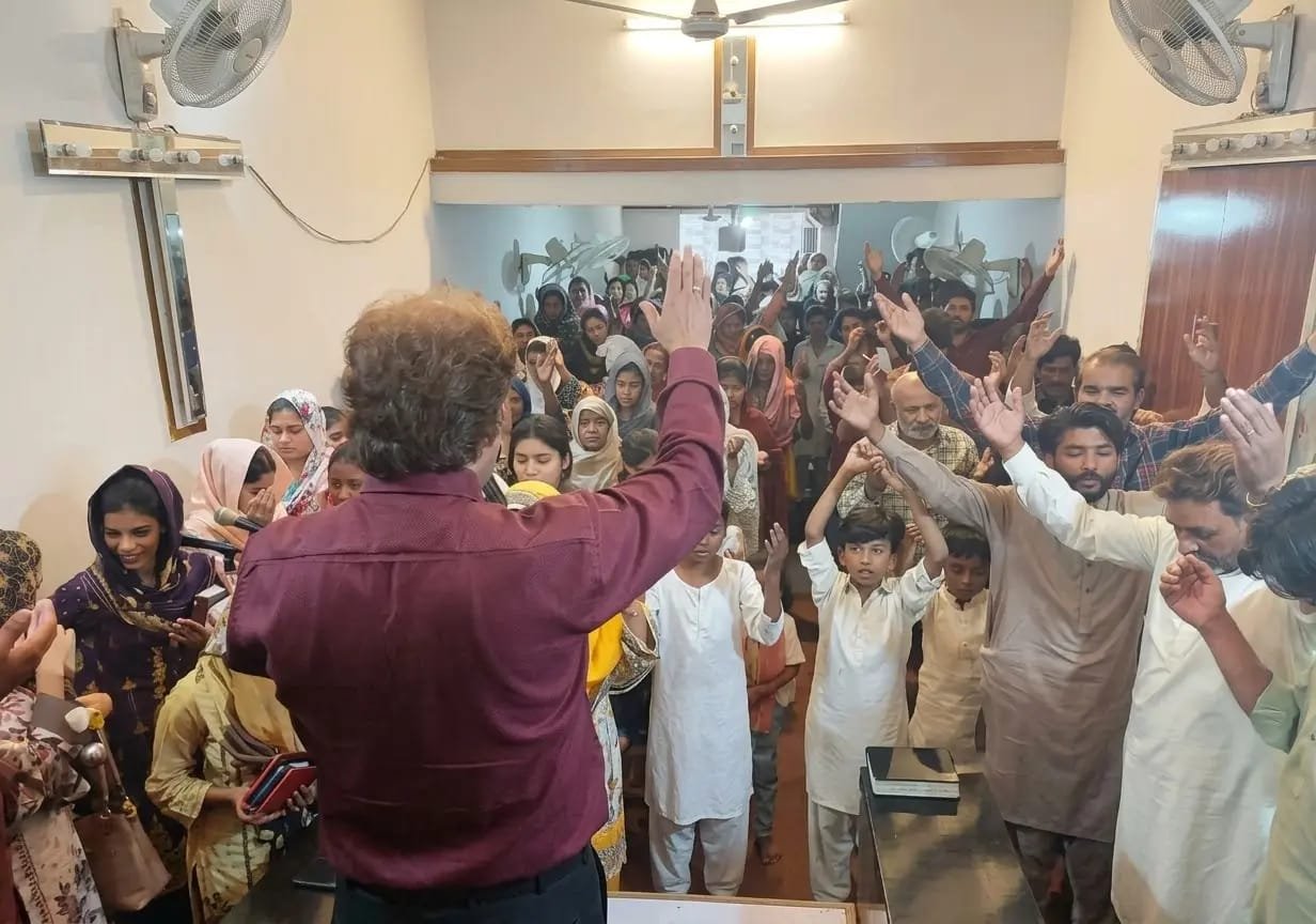 Pastor Yousaf Hussain leading worship with congregation raising hands at Church of Christ Assembly in Pakistan